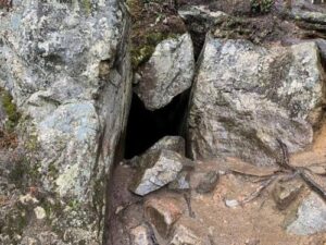 Una foto dell'ingresso della grotta della Chiesa del Diavolo. È una fessura stretta tra le rocce che forma una sorta di forma a clessidra sul lato. Le rocce circostanti sono nude con muschio sparsi su di esse e il pavimento fuori dalla grotta è composto da terra fine e pezzi di pietra.