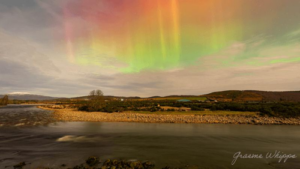 una foto di un fiume e della campagna scozzese dietro di esso e sopra le luci del nord che brillano