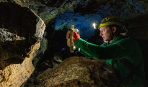 L'astronauta della NASA Jessica Wittner si trova all'interno di una grotta di lava tenendo in mano una nuova fotocamera lunare prototipo e scattando una foto di una roccia. Illumina la roccia con una torcia frontale.