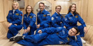 six-people-in-blue-space-suits-in-front-of-a-wooden-wall-with-a-hatch-behind-them-one-man-laying-down-in-front-of-the-others-who-are-squatting-in-a-paint-me-like-your-french-girls-pose-m.png