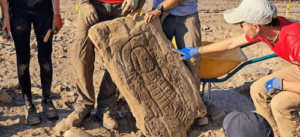 Una foto degli archeologi che tengono in mano la lastra di pietra che formava la stele funeraria. La foto è ritagliata in modo da poter vedere solo tre coppie di gambe accovacciate intorno alla pietra - che arriva all'altezza dell'anca - e un individuo con un cappello bianco e una maglietta rossa che si china per indicare le incisioni.