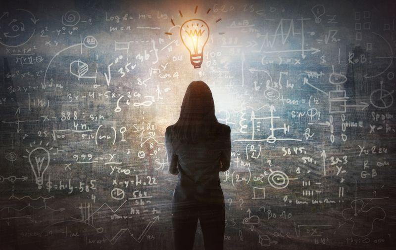 person-with-long-hair-in-business-suit-facing-away-from-the-camera-towards-blackboard-with-chalk-equations-on-illustrated-lightbulb-above-their-head-m-1.jpg