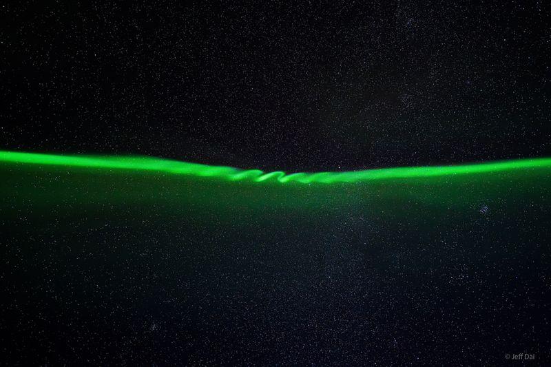 L'aurora boreale si vede dall'alto, una sottile striscia di luce verde contro il cielo notturno. Al centro si possono vedere una serie di onde