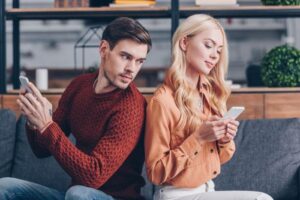 Uomo dai capelli castani con maglione rosso che guarda sospettosamente il telefono di una donna bionda in una camicia marrone