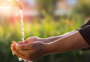 Le mani di un bambino indiano raccolgono acqua mentre scorre da un rubinetto esterno. L'immagine è fortemente saturata dalla luce solare che sfoca l'angolo in alto a sinistra dove si trova il rubinetto. Lo sfondo sfocato mostra un giardino verde lussureggiante o uno spazio aperto simile.