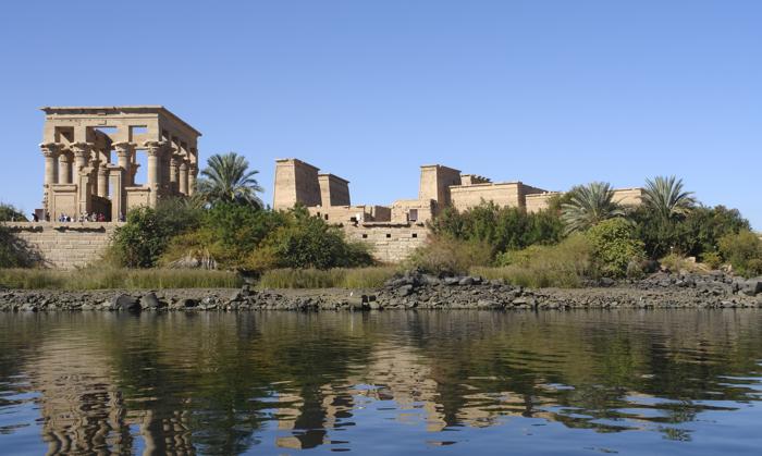 Una foto del Tempio di Philae scattata sul Nilo. Il primo piano mostra la superficie dell'acqua, poi un argine roccioso seguito da una fila di erbe e alberi. Dietro di essi si erge il tempio in arenaria chiara. A sinistra c'è un insieme di archi rialzati, e poi al centro dell'immagine si trova un complesso più compatto.