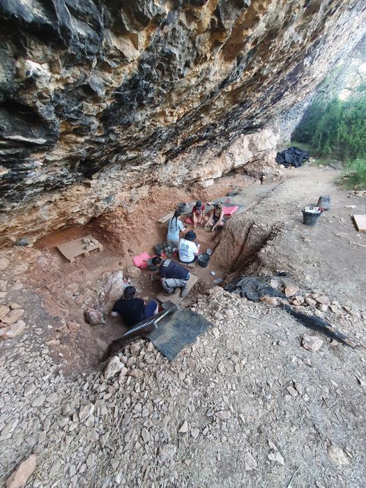 ricercatori che scavano presso Abric Pizarro, ripresi dall'alto; sono seduti in una trincea poco profonda alla base di una parete rocciosa a strapiombo