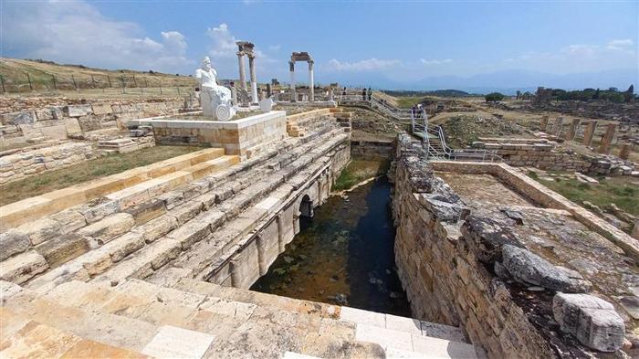 Una foto delle rovine della città greco-romana di Hierapolis, che si trova nella Turchia moderna. La foto mostra un canale tra un muro e una struttura a gradini che potrebbe essere l'ingresso all'arena del cancello di Pluto. Sopra l'arco nel canale c'è una statua che potrebbe raffigurare il dio ctonio stesso.