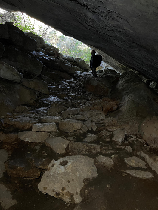 ingresso diagonale di una grotta con una persona in controluce contro gli alberi e il cielo, parte del sentiero roccioso che scende nella grotta è visibile verso il fotografo