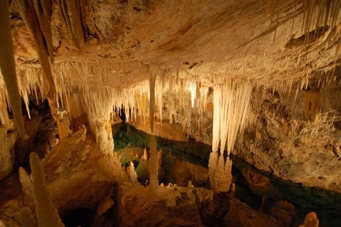 Grande caverna sotterranea piena di stalattiti e stalagmiti con acqua blu brillante sul fondo.