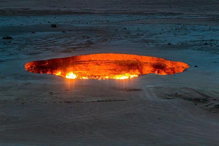 Una foto del cratere di Darvaza scattata da lontano. Lo scatto mostra il grande cratere da una posizione elevata nel deserto aperto. La foto è stata scattata dopo il tramonto, quindi l'intero buco sembra brillare di fiamme.