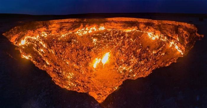 Una foto scattata di notte del cratere di Darvaza, che mostra uno spazio grande a forma di triangolo rovesciato che brilla di fuoco. Ci sono zone del cratere più luminose di altre, mentre il bordo superiore del buco ha una linea di fuoco quasi ininterrotta che lo circonda.