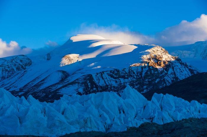 La foto mostra le cime delle montagne e il ghiaccio intorno all'altopiano tibetano. In primo piano ci sono formazioni di ghiaccio frastagliate dietro le quali è visibile una superficie rocciosa che è poi coperta dalla neve. La maggior parte dello scatto è stata scattata all'ombra, ma ci sono rigonfiamenti nella neve lontana che sono illuminati dalla morbida luce del mattino.