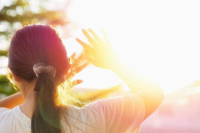 Persona con capelli scuri raccolti in una coda che tiene le mani alzate per proteggersi dal sole accecante