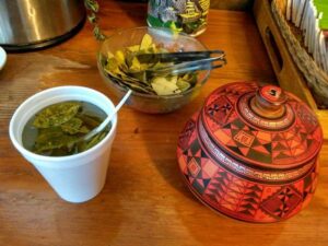 photograph-left-to-right-of-a-polystyrene-cup-of-coca-leaf-tea-a-glass-bowl-of-coca-leaves-with-a-pair-of-metal-tongs-and-an-orange-red-patterned-bowl-with-a-lid-m.jpg