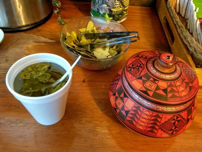 photograph-left-to-right-of-a-polystyrene-cup-of-coca-leaf-tea-a-glass-bowl-of-coca-leaves-with-a-pair-of-metal-tongs-and-an-orange-red-patterned-bowl-with-a-lid-m.jpg