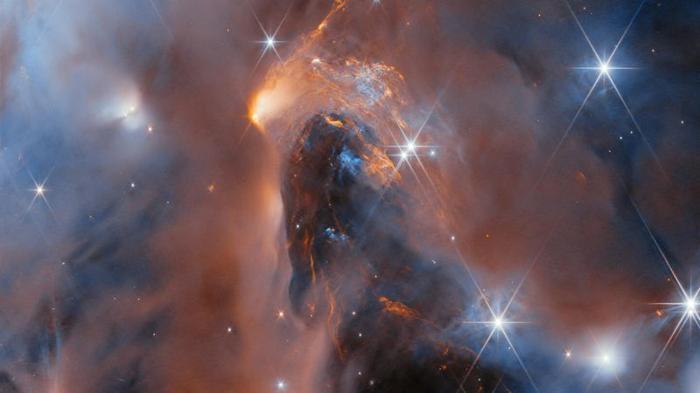 Whispy dark clouds are intersped with bright glowing ones, together with bright stars and smaller dot-like object in this panorama of a nebula.