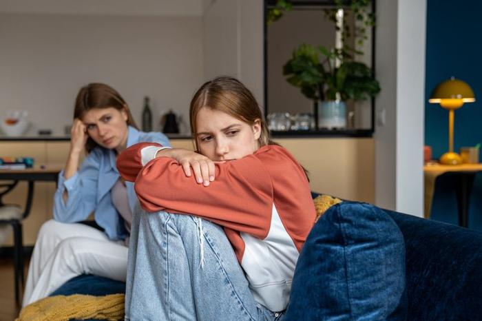 La foto mostra una ragazza adolescente seduta su un divano con sua madre sullo sfondo. La ragazza è seduta di profilo mentre guarda verso la fotocamera. Ha le ginocchia tirate verso il corpo quindi sembra contrariata mentre sua madre è seduta all'altro capo del divano e la guarda con uno sguardo preoccupato o frustrato. La scena è ambientata in un soggiorno.