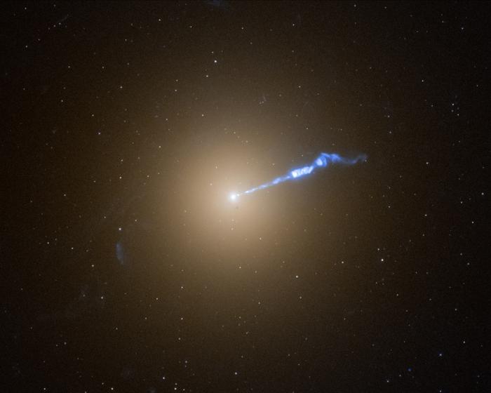 Una foto di Hubble della galassia M87, che assomiglia a un batuffolo di cotone bianco traslucido e sfocato. La luminosità diminuisce gradualmente in tutte le direzioni da un punto luminoso bianco al centro. Un jet ondulato di materiale blu-bianco si estende dal nucleo a forma di punto verso l'esterno fino all'angolo superiore destro, circa a metà della galassia. Le stelle punteggiano lo sfondo