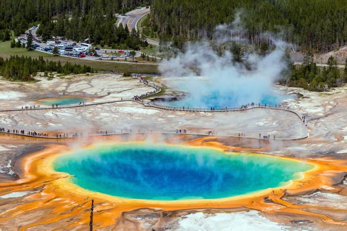 Una foto aerea della Grand Prismatic Spring di Yellowstone che mostra il blu intenso della sorgente stessa, così come i suoli gialli e arancioni circostanti macchiati. Dall'acqua si alza del vapore. Sullo sfondo, un'altra sorgente è visibile, quasi completamente coperta da spesse colonne di vapore. I pontili sono anche visibili serpeggiando intorno alle sorgenti mentre i turisti le visitano.