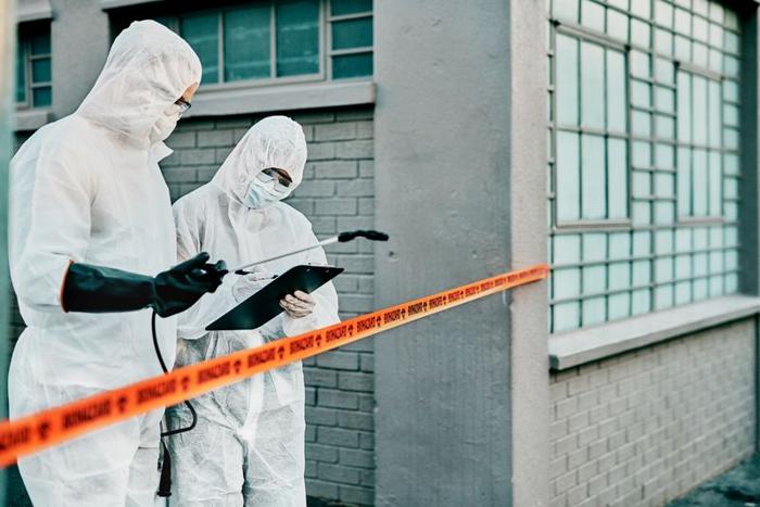 due persone in tute HazMat che guardano una clipboard, in piedi dietro al nastro arancione di biohazard