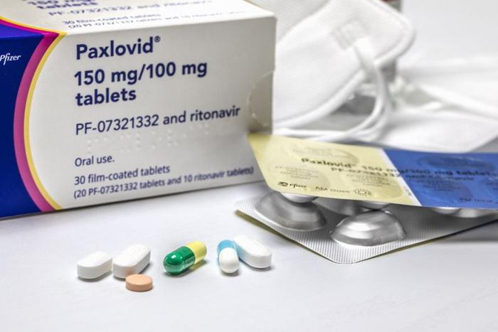 Paxlovid box on a table, with some open blister packs and some N95 masks in the background, and a selection of different coloured pills and capsules in the foreground.