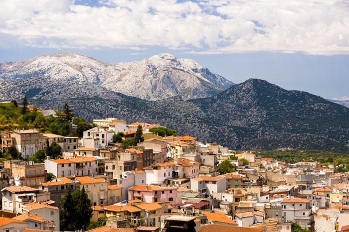 Una foto di una città in Sardegna, Italia incastonata su un pendio. I tetti di argilla rossa possono essere visti inclinarsi da sinistra a destra dell'immagine. Gli edifici sono tutti bagnati di luce solare. Sullo sfondo sono visibili montagne innevate sotto un cielo parzialmente nuvoloso.