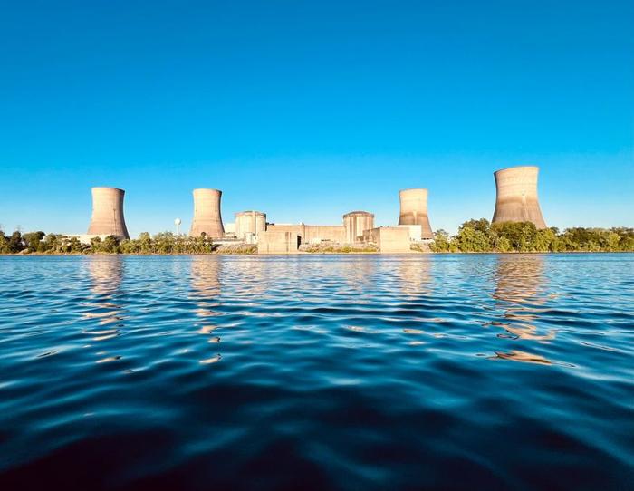 Una foto scattata dall'altra parte dell'acqua che mostra la centrale elettrica di Three Mile Island. Le quattro torri di raffreddamento della centrale sono visibili in quanto sono raggruppate intorno agli edifici del reattore principale. La foto è stata scattata in una giornata luminosa e il cielo dietro la centrale è completamente blu.