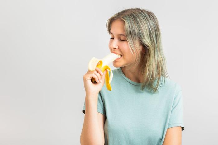 donna su sfondo beige indossa una maglietta verde menta con capelli biondi che morde una banana