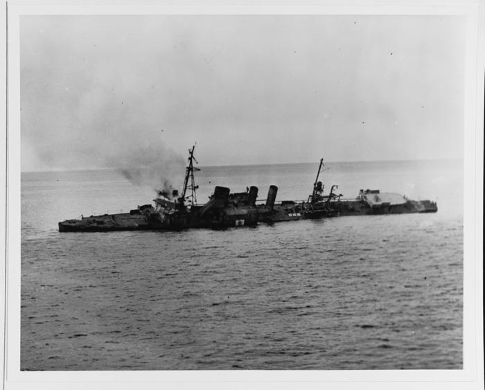 Una foto in bianco e nero che mostra l'USS Stewart mentre affondava sotto l'acqua. La nave si è inclinata verso la fotocamera sul lato sinistro e c'è una colonna di fumo che si alza vicino alla prua. 