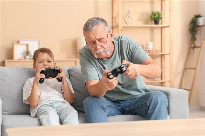 La foto mostra un anziano uomo nonno che gioca a un gioco Playstation con il suo giovane ragazzo, presumibilmente suo nipote. I due sono seduti su un divano grigio di pietra in un soggiorno. L'uomo, che indossa gli occhiali e ha una barba senza mento, si sta inclinando leggermente verso destra mentre gioca.