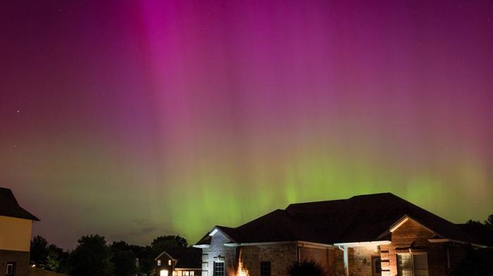 alcune case sono visibili in fondo alla foto. Nel cielo notturno sono visibili una striscia verde e una striscia viola, l'aurora