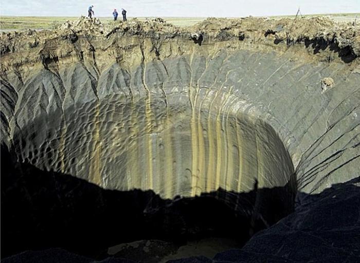 Una foto mostra l'enorme cratere causato dalle esplosioni. Il buco ha un bordo di argilla grigia che poi scende in un profondo pozzo largo decine di piedi. Sullo sfondo dell'immagine, tre persone stanno guardando nel buco ma sono minuscole rispetto alla sua dimensione.