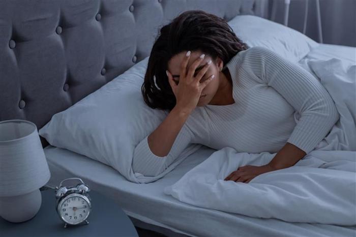 Una foto di una donna che indossa una camicia da notte bianca che si tiene il viso mentre è sdraiata sul letto. L'orologio sul comodino mostra che sono le 6:15