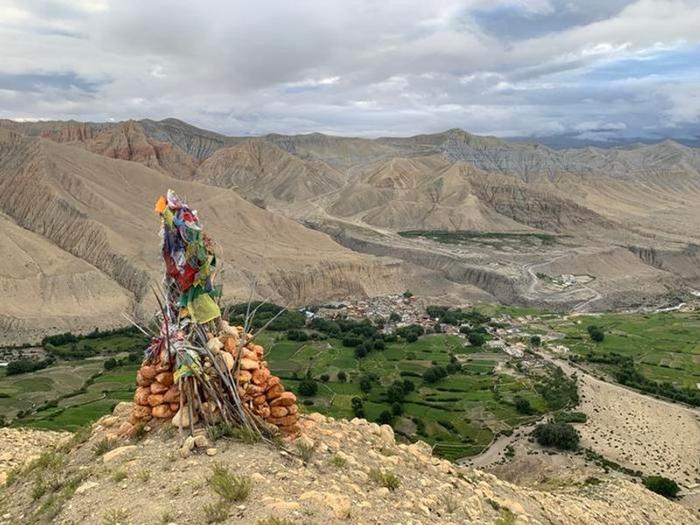 Una foto che mostra un monumento fatto di pietre rosse di argilla ammucchiate e sottili bastoncini assemblati intorno a quello che sembra essere bandiere di colori diversi. Il monumento si trova su un pendio che guarda giù su un villaggio tibetano incastonato tra campi verdi tra pendii aridi e aspri.