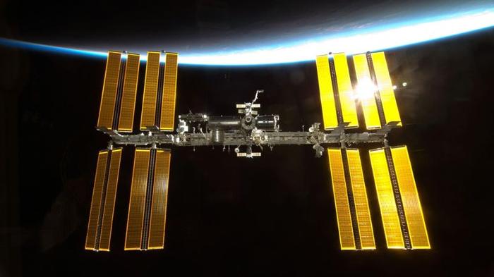 La stazione spaziale internazionale è al centro dell'immagine. Il Sole fa sembrare dorati e lucenti i pannelli solari. La Terra è appena illuminata sopra la ISS