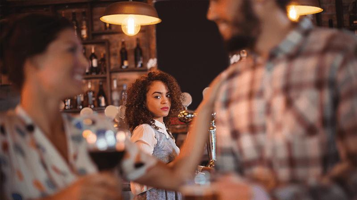 Due persone che flirtano in un bar, una donna sullo sfondo che sembra gelosa.