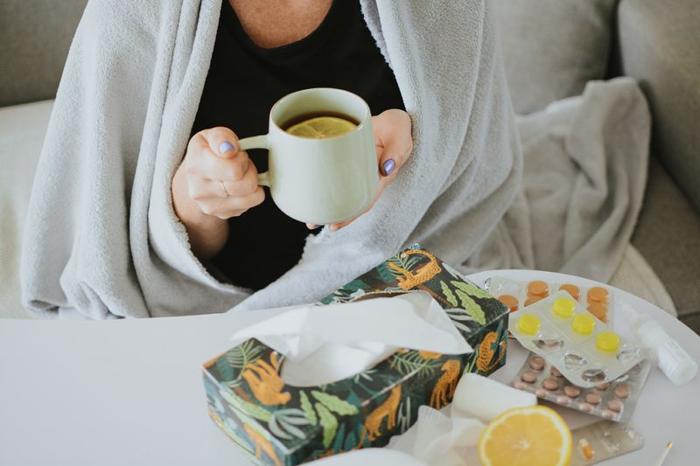 una donna beve tè con una fetta di limone mentre è avvolta in una coperta (solo il suo torso è visibile); sul tavolo di fronte a lei c'è una scatola di fazzoletti, una selezione di pastiglie e pillole, uno spray nasale e il resto del limone