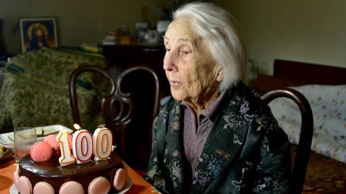 anziana donna seduta a un tavolo con una torta di compleanno, con candele a forma di numero 100