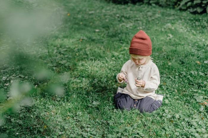 Un piccolo ragazzo biondo con un berretto siede fuori sull'erba.