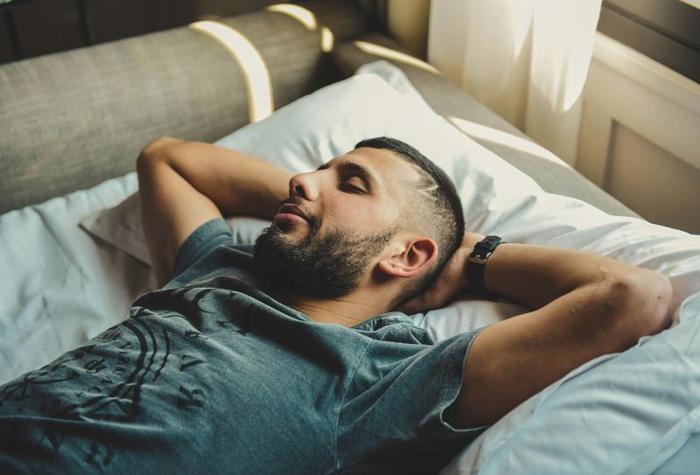 uomo sdraiato sulla schiena con le braccia dietro la testa su un letto, che dorme; indossa una maglietta verde e le coperte del letto sono bianche