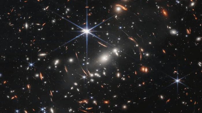 Al centro dell'immagine, tra le 4 e le 6 in punto negli sprazzi della stella luminosa, ci sono diverse galassie luminose e bianche. Queste sono membri del cluster di galassie. Lo sfondo dello spazio è nero. Migliaia di piccole galassie appaiono nell'immagine. I loro colori variano. Alcune sono tonalità di arancione, altre sono bianche. La maggior parte appare come ovali sfocate, ma alcune hanno braccia spiraliformi distintive. Ci sono anche molti sottili, lunghi archi arancioni. Seguono cerchi concentrici invisibili che si curvano intorno al centro dell'immagine. Queste sono immagini di galassie di sfondo che sono state allungate e distorte dal cluster di galassie in primo piano.