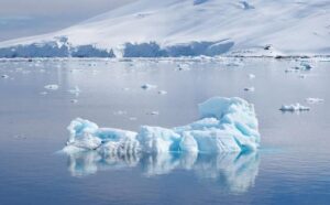 La foto mostra un pezzo di ghiaccio galleggiante in un corpo d'acqua immobile con una montagna coperta di neve alle spalle. Il ghiaccio ha una sfumatura bluastra e ci sono altri pezzi di ghiaccio sparsi per l'acqua.