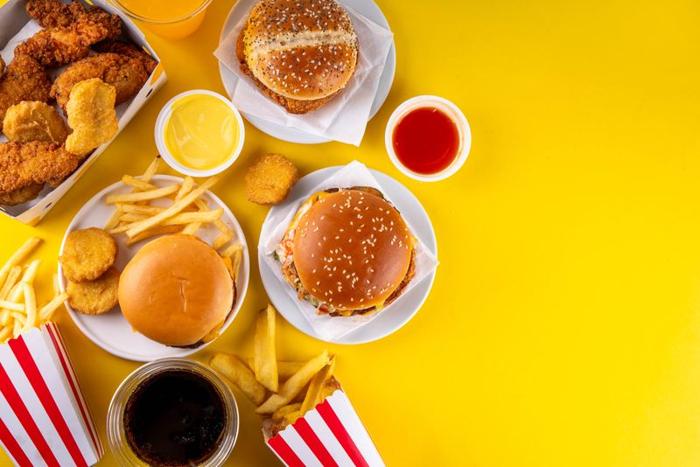 La vista dall'alto del cibo su una superficie gialla. Il cibo è stato raccolto a sinistra dello scatto e mostra tre hamburger - uno con un panino semplice - circondati da patatine fritte e nuggets di pollo. Ci sono anche alcune ciotole con salse di diversi colori. Le patatine sono servite in scatole a righe rosse e bianche.