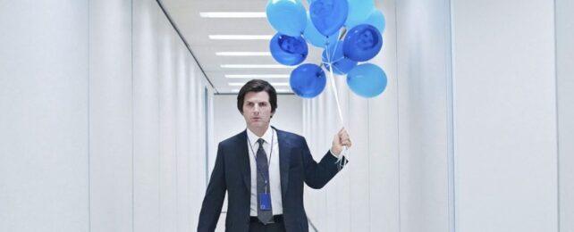 Attore Adam Scott in un completo da lavoro che tiene palloncini blu in un lungo corridoio bianco d'ufficio