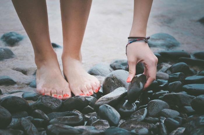 Una donna con unghie dei piedi e delle mani dipinte sta in piedi su una spiaggia rocciosa, raccogliendo sassi.