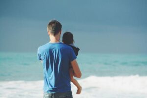 uomo che tiene un bambino in braccio, entrambi di spalle rispetto alla telecamera mentre guardano l'oceano aperto