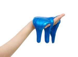 Un primo piano della mano di una persona che tiene della slime blu nella mano. La slime sta colando sul palmo e sta iniziando a cadere a terra. La mano è impostata su uno sfondo bianco.